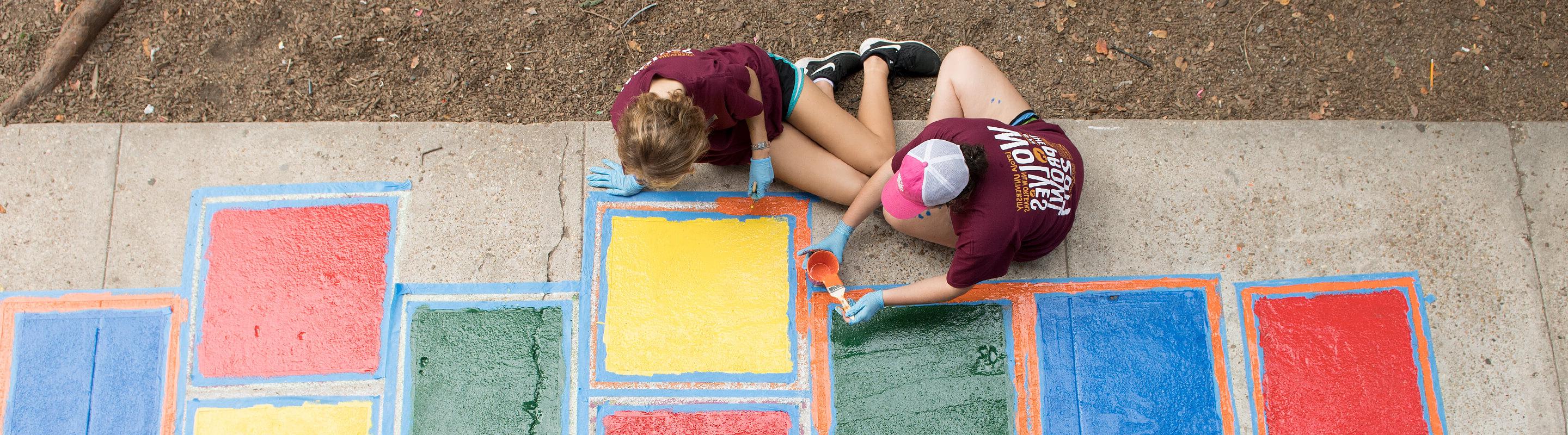 Two alumni painting hopscotch on a sidewalk 